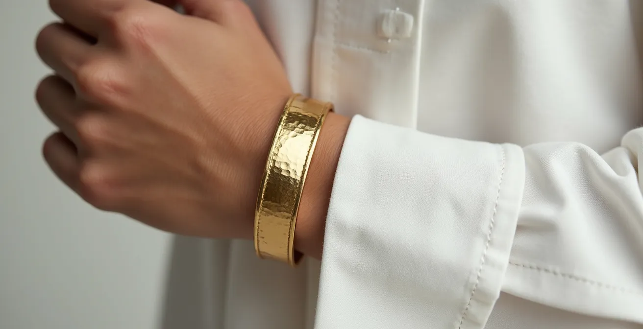Nahaufnahme von minimalistischem Outfit mit einzelnem auffälligem Accessoire wie einem gehämmerten Goldarmreif an einem weißen Leinenhemd.